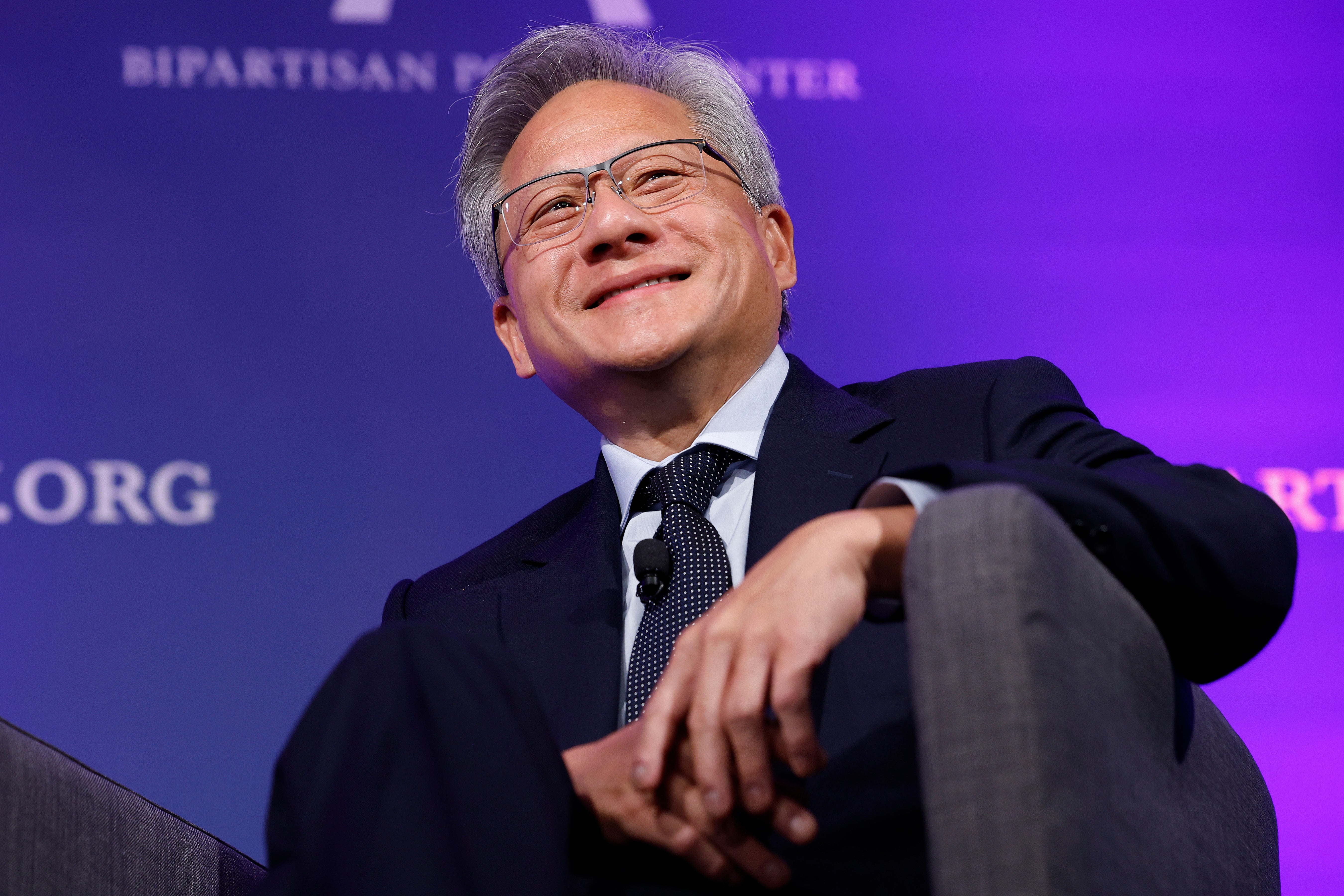 Jensen Huang sits on a chair with folded hands, smiling, against a purple background